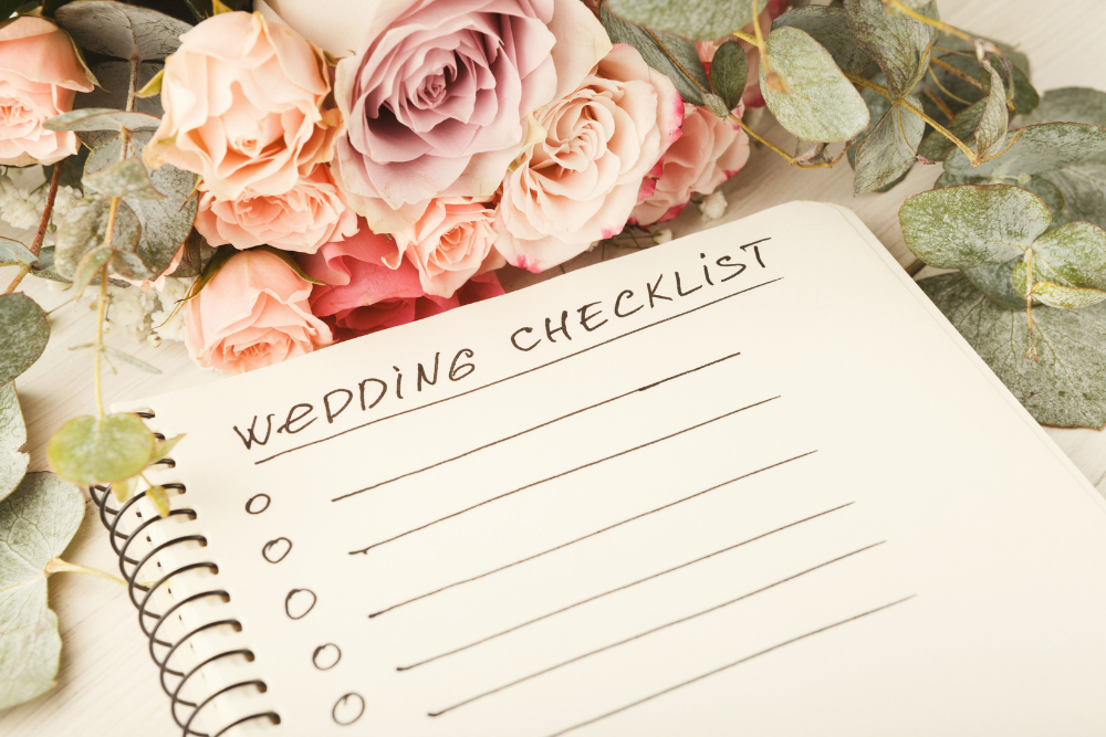 Open notebook with wedding venue checklist surrounded by pink roses and greenery, symbolizing essential planning for couples.